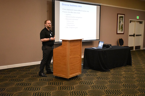 A man speaks from behind a podium. A projector screen is beside him displaying text “Diverse business rules.” A laptop is open and plugged into a projector on a table.
