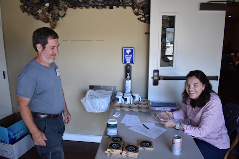 A smiling woman seated at a table to the right. A man stands in front of the table on the left.